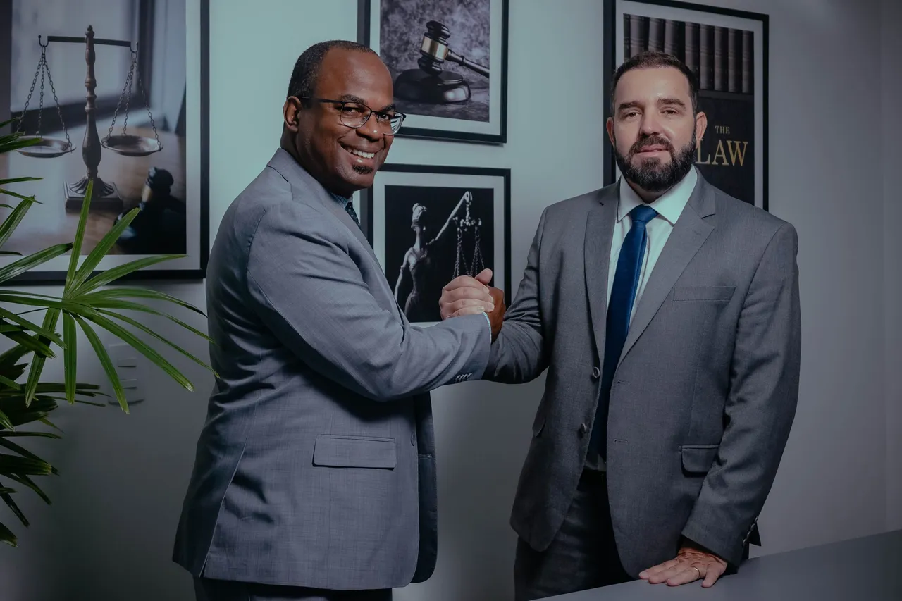 Dr. Rodrigo Higino e Dr. Luiz Otávio Franco, de terno cinza, se cumprimentando no escritório, com quadros de balança, martelo de juiz e estátua da justiça ao fundo.